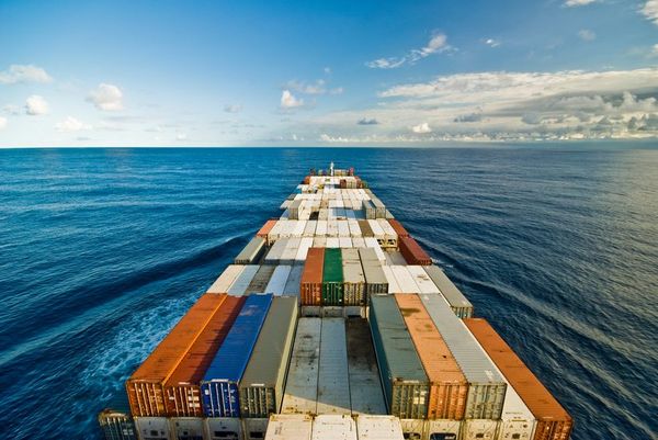 A large cargo ship loaded with multicolored shipping containers sails across a vast blue ocean under a cloudy sky