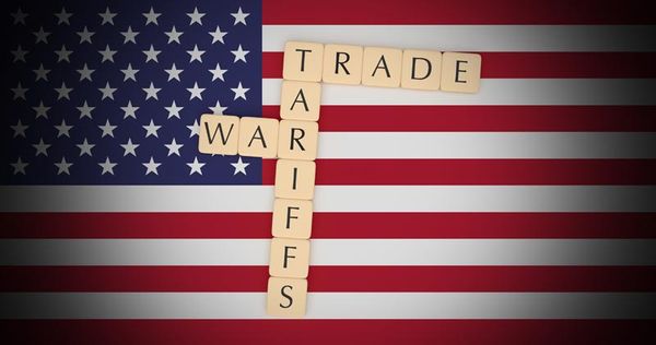 Scrabble tiles spelling "TRADE" and "TARIFFS" vertically against a blurred American flag background, suggesting