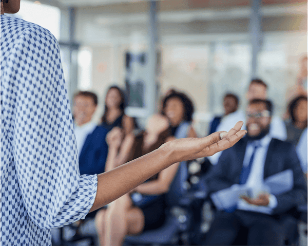 A person in a blue and white patterned shirt gestures while presenting to a blurred audience
