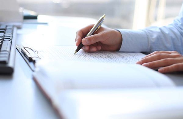 Hand in blue shirt sleeve writing with a silver pen on lined paper near a keyboard and clipboard