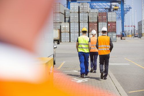 Ree workers in safety vests and hard hats walk away from the camera in a shipping container yard with stacked