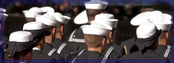 Navy sailors in dress whites stand in formation, backs turned, white caps crisp against dark uniforms with striped sleeves
