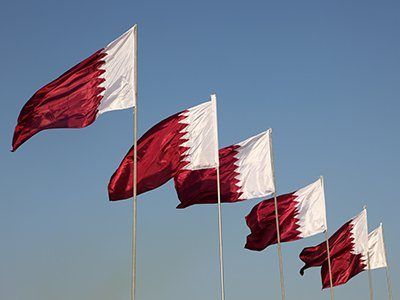 Five Qatari flags of decreasing size, waving against a clear blue sky featuring red and white horizontal stripes