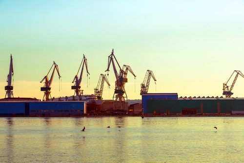 Industrial port scene with tall silhouetted cargo cranes casting long shadows over calm water at sunset or sunrise